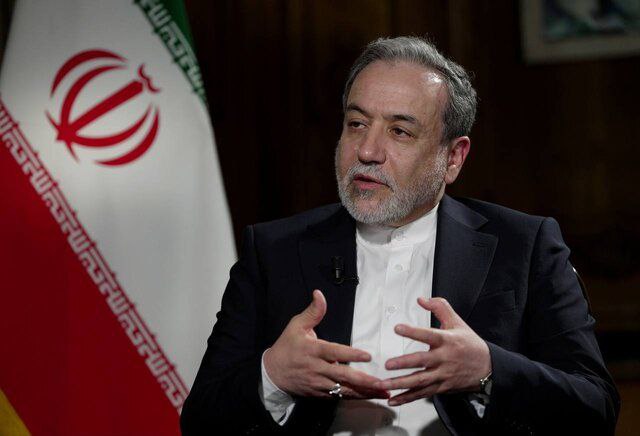 Iranian official discussing diplomatic issues, seated in front of the Iranian flag, emphasizing key points with hand gestures during an interview.