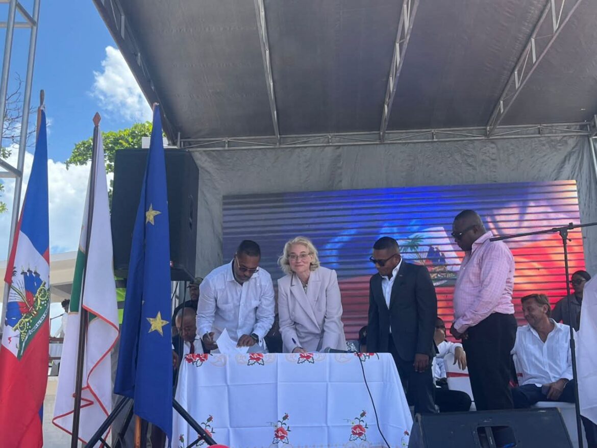 Officials signing a document during a formal event, with flags of Haiti and the European Union displayed, under a tent with a backdrop of vibrant colors.