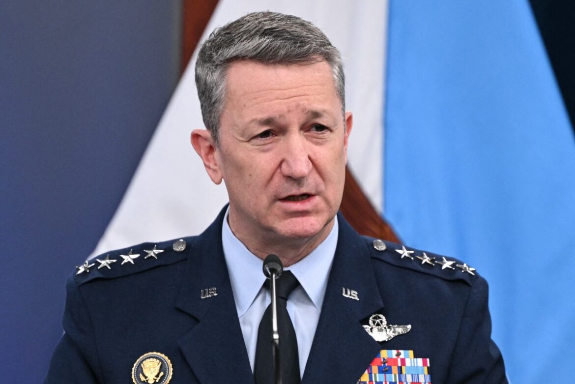 U.S. Air Force General speaking at a press conference, wearing a military uniform with various medals and insignia, set against a backdrop of flags.