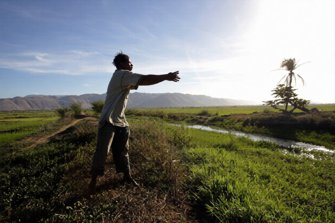 A man standing barefoot on a grassy hillside extends his arms towards the sky, surrounded by lush green fields and mountains in the background, under a bright blue sky.