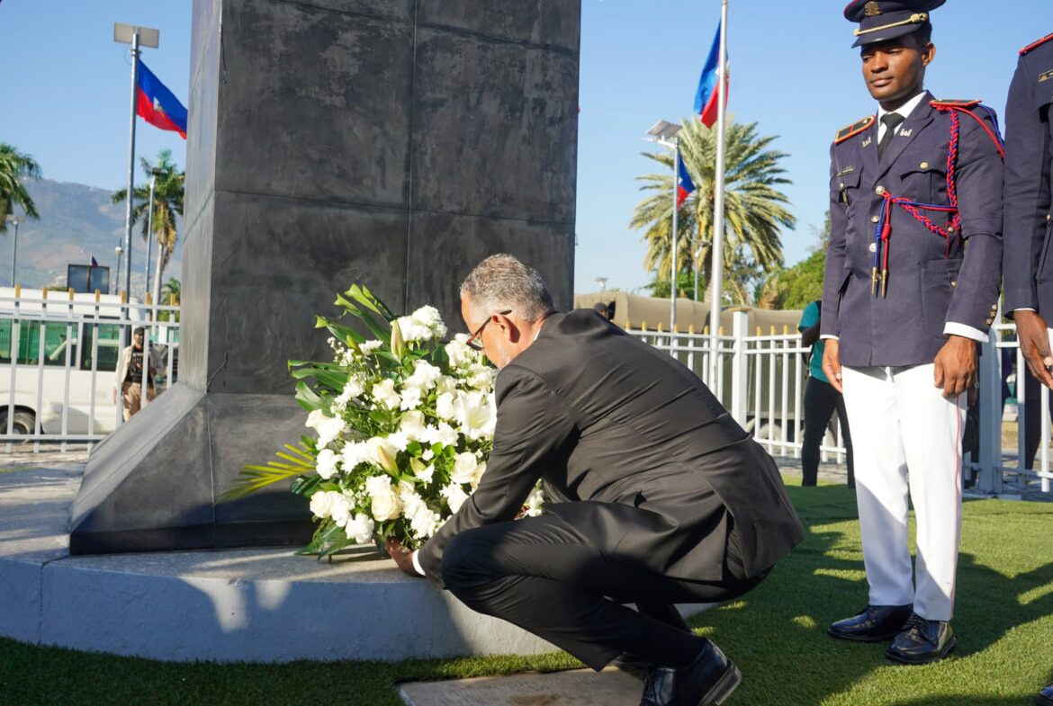 A man in a black suit kneels to place a bouquet of white flowers at a memorial, surrounded by flags and uniformed officers in a ceremonial setting.