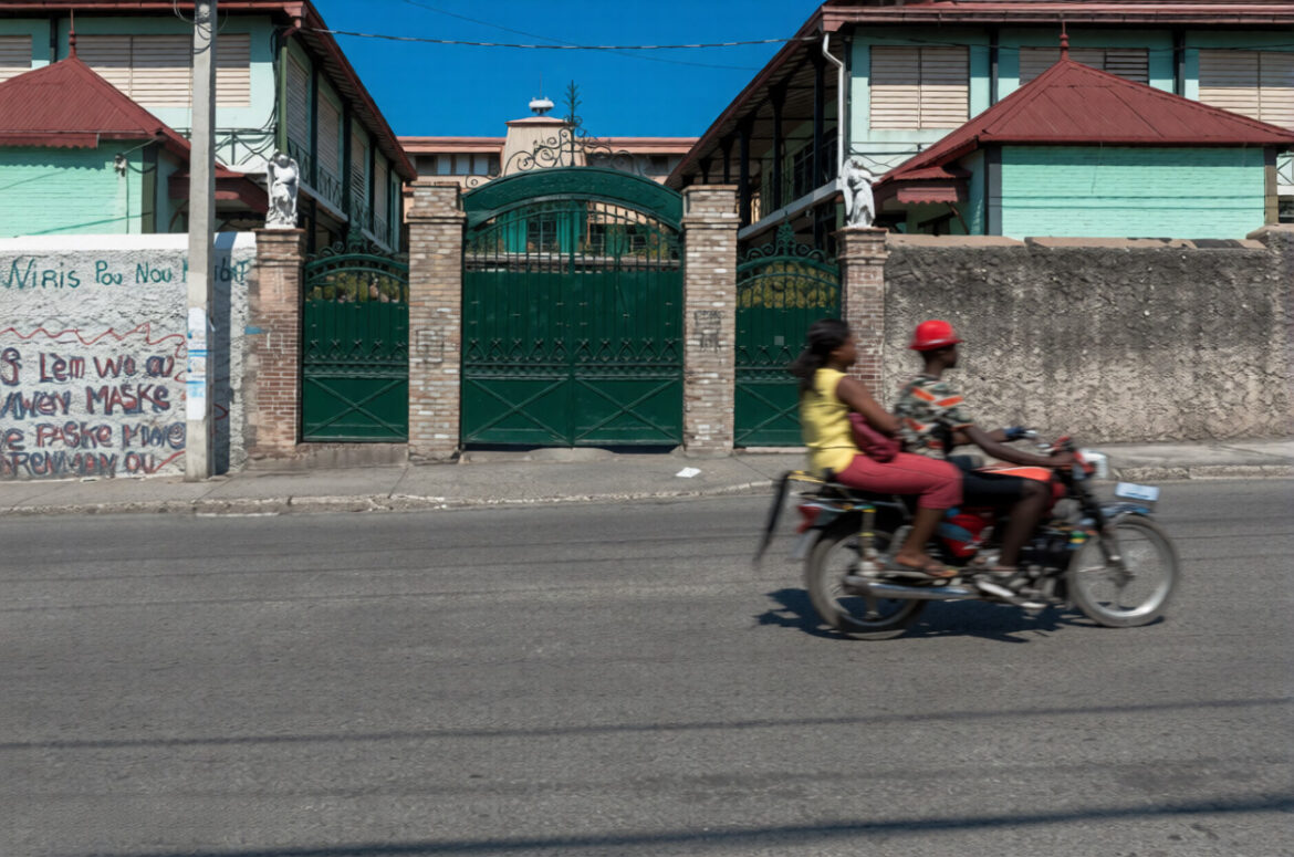 Two individuals riding a motorcycle pass by a green gate in front of a residential building with colorful facades under a clear blue sky.