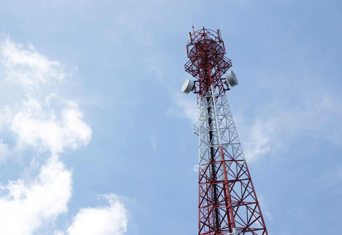 A tall telecommunications tower with a red and white structure, equipped with antennas and satellite dishes, against a clear blue sky with scattered clouds.