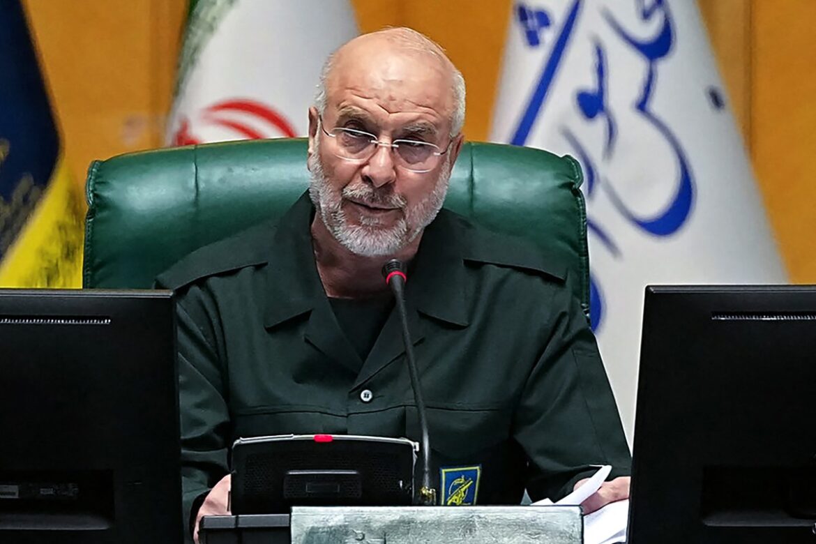 Iranian political figure speaking at a conference, wearing a dark green military-style uniform, with a serious expression. Flags and official symbols are visible in the background, indicating a formal setting.