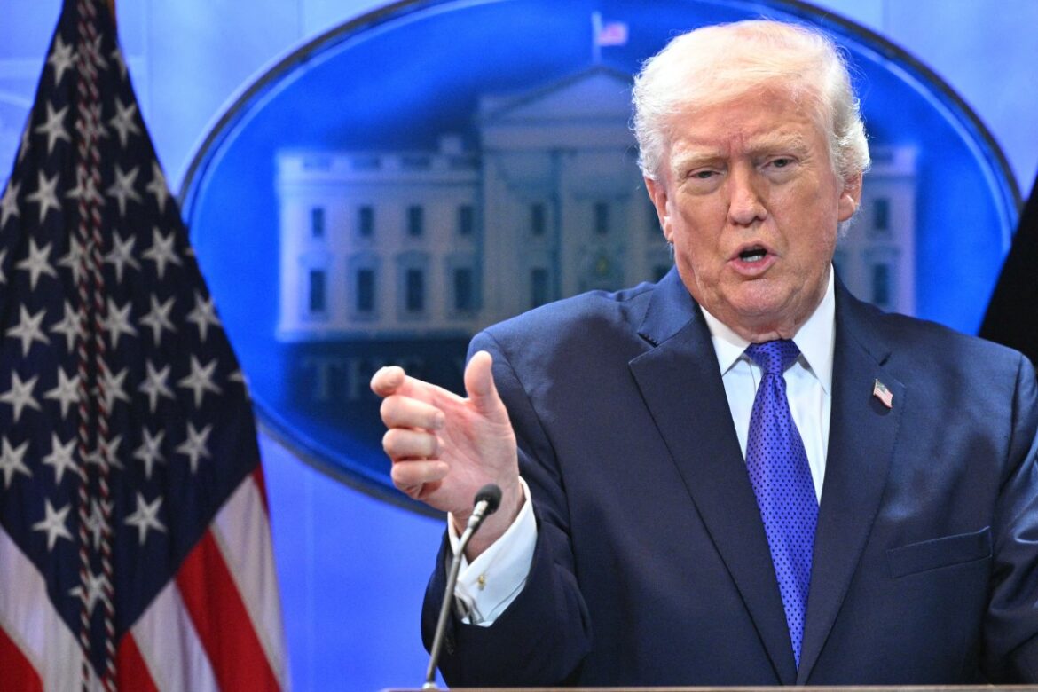 Former President Donald Trump speaking at a press briefing, gesturing with his hand, with the White House visible in the background and an American flag to his side.