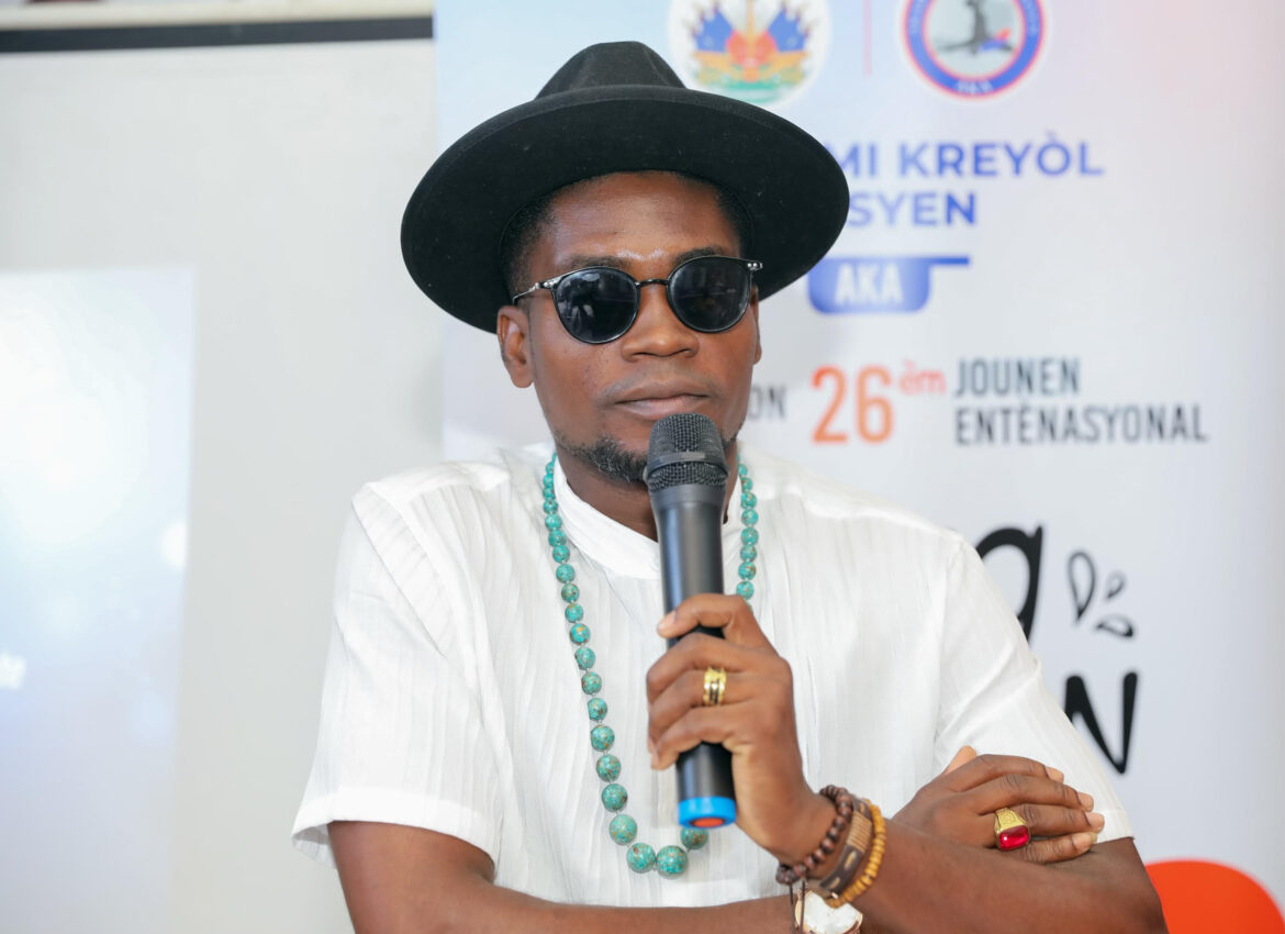 A man wearing sunglasses, a wide-brimmed hat, and a white shirt speaks into a microphone at an event promoting Haitian Creole culture, with a banner in the background displaying the 26th International Creole Day.