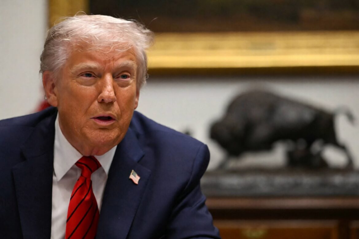 Close-up of a man in a suit with a red tie, seated at a table, speaking during a meeting. A bronze sculpture of a buffalo is visible in the background, along with a decorative frame.