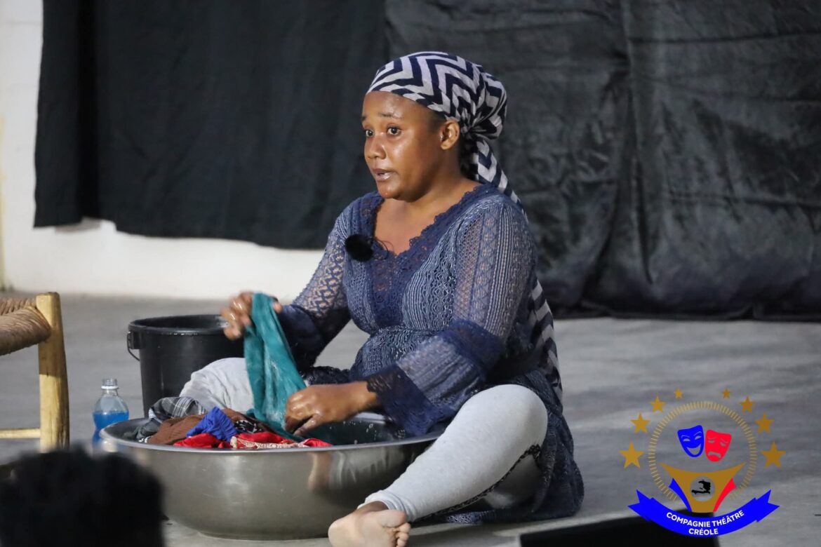 A woman performing a dramatic scene while washing clothes in a bowl, showcasing expressive emotions. She wears a blue lace top and a striped headscarf, with a black backdrop enhancing the theatrical setting. The image highlights the art of storytelling through everyday activities in a cultural performance.
