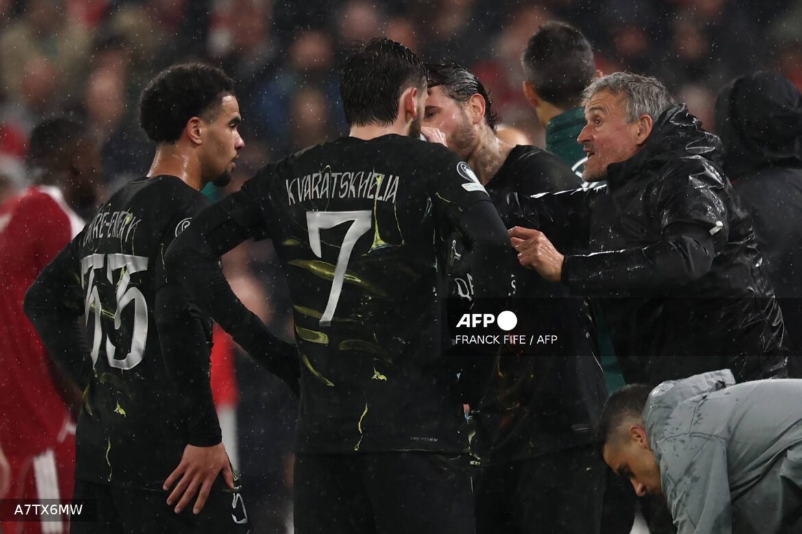 Football players in black jerseys, including Kvaratskhelia, gather during a rain-soaked match, engaging in a heated discussion with their coach, who is wearing a black rain jacket. The scene captures the intensity of the game and the team's dynamics.