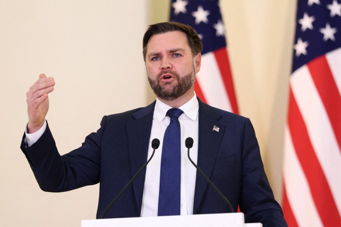 Man in a suit delivering a speech at a podium with American flags in the background, emphasizing key points with hand gestures.