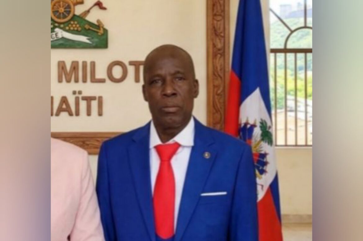 Official portrait of a Haitian politician in a blue suit with a red tie, standing in front of the Haitian flag and a wall featuring the words "MILON" and "HAÏTI."