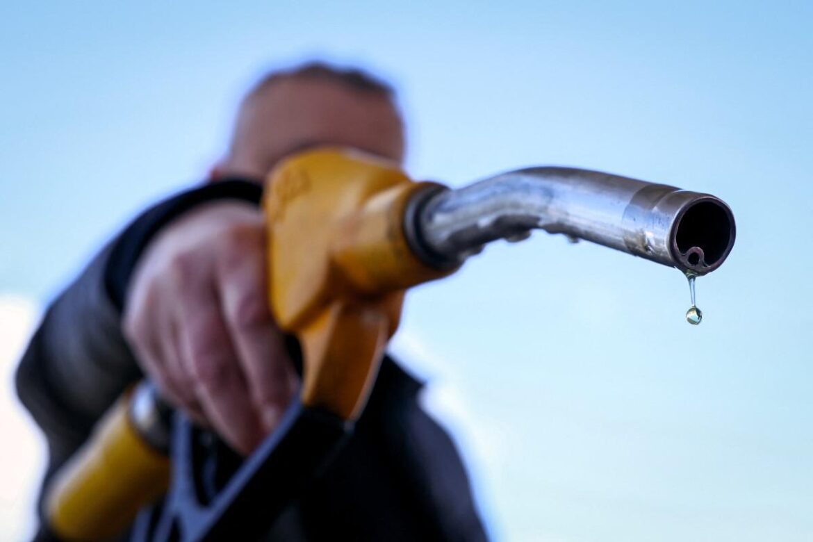 Close-up of a fuel nozzle with a droplet of gasoline, held by a person at a gas station against a clear blue sky, illustrating fuel dispensing.
