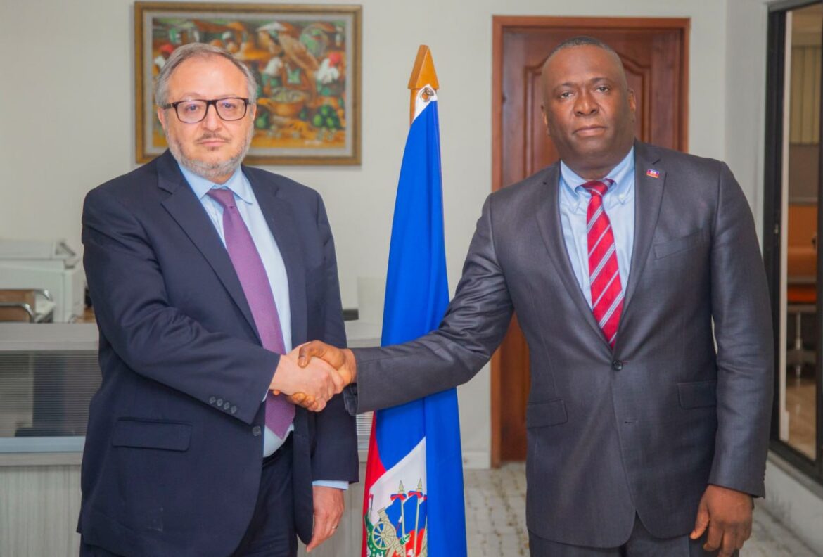 Two men shaking hands in an office setting, with a Haitian flag and a colorful painting in the background. The first man, wearing glasses and a suit, represents a diplomatic or business context, while the second man, dressed in formal attire, appears to be a local official. This image symbolizes collaboration and partnership.