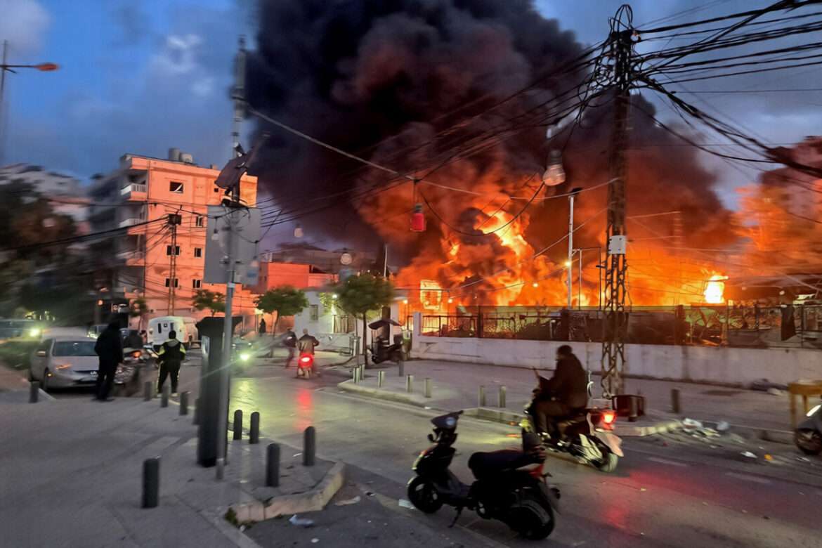 Fire and thick black smoke billow from a building in an urban area, with emergency response vehicles and individuals on motorcycles navigating the street. Power lines and streetlights are visible, highlighting the chaotic scene during dusk.