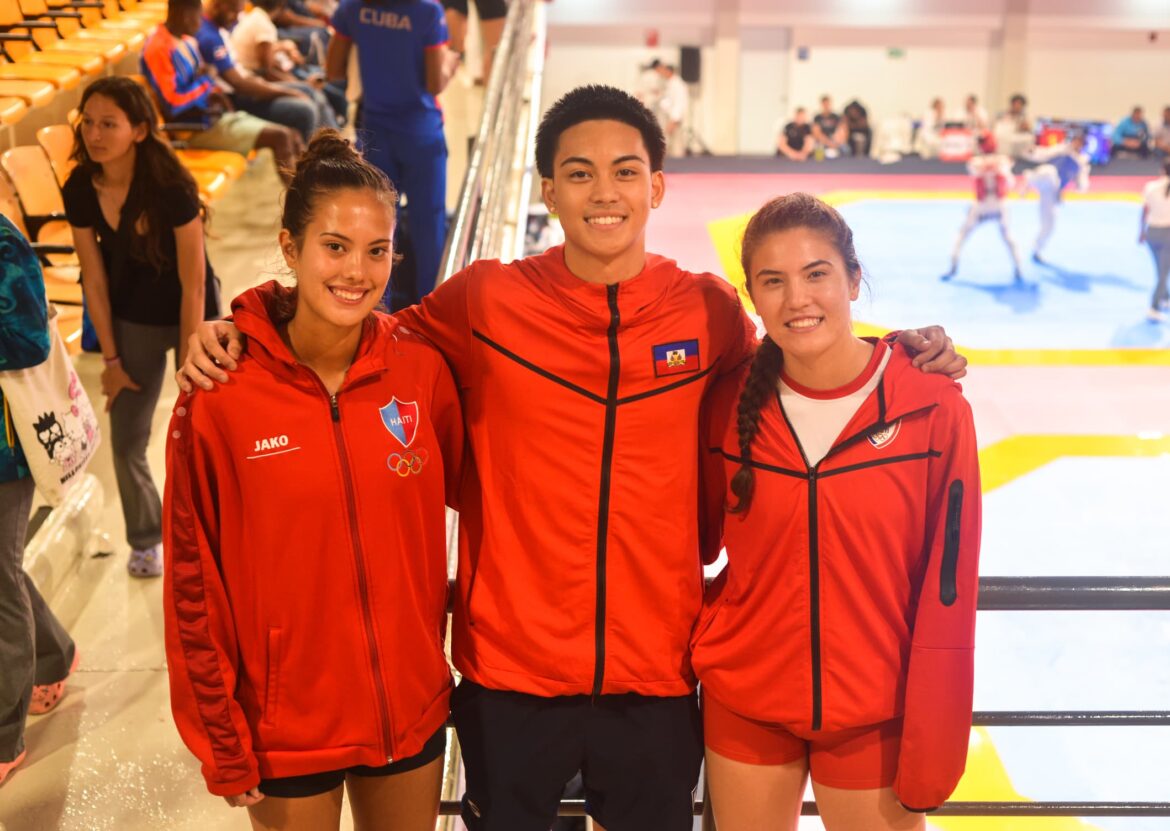 Three athletes in red jackets pose together at a sports venue, showcasing team spirit. The background features a martial arts competition in progress, highlighting an energetic atmosphere.